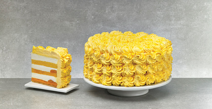 A cake yellow buttercream swirl frosting next to a slice of the same cake with orange to yellow ombre cake layers against a gray backdrop. 