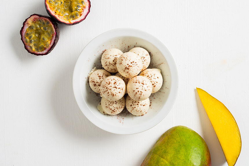 white passionfruit-mango truffles in a bowl in the center of the image, with cut open passionfruit at the top left corner of the image, and sliced mango in the bottom right corner of the image. 