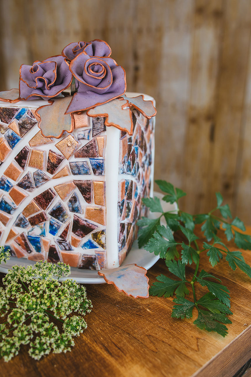 A large cube cake sits on a plate on a wooden table. The cake is decorated with edible mosaic tiles and topped with purple fondant roses and gray fondant leaves, edged with copper brilliant powder. Leaves and white flowers sit in front of the cake, with wooden boards in the background. 
