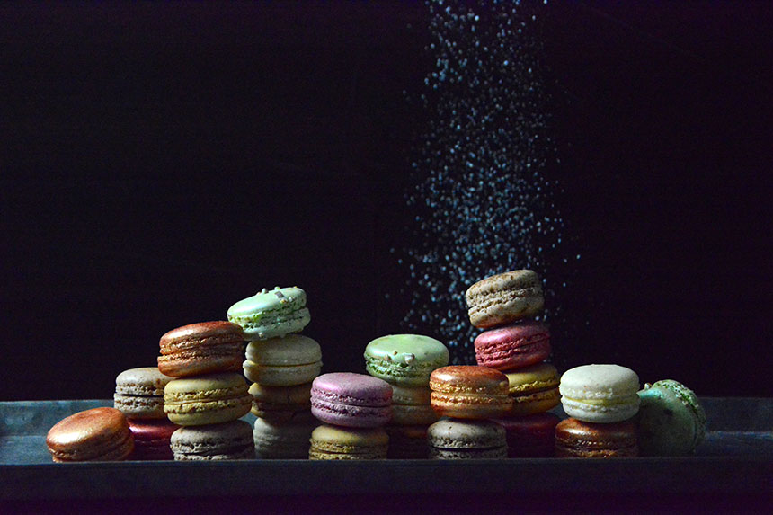 Shiny, multicolored macarons sit against a dark background with brilliant powder being sprinkled on top. 
