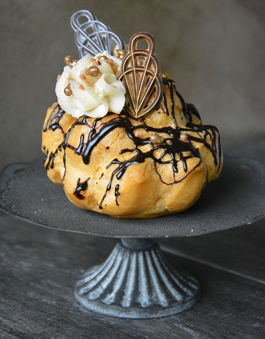 An image of a cream puff topped with cream, crispearls and chocolate decor. 