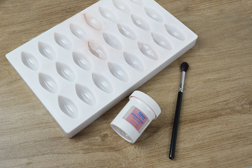 A white bonbon mold sitting text to a container of brilliant powder and a brush. 