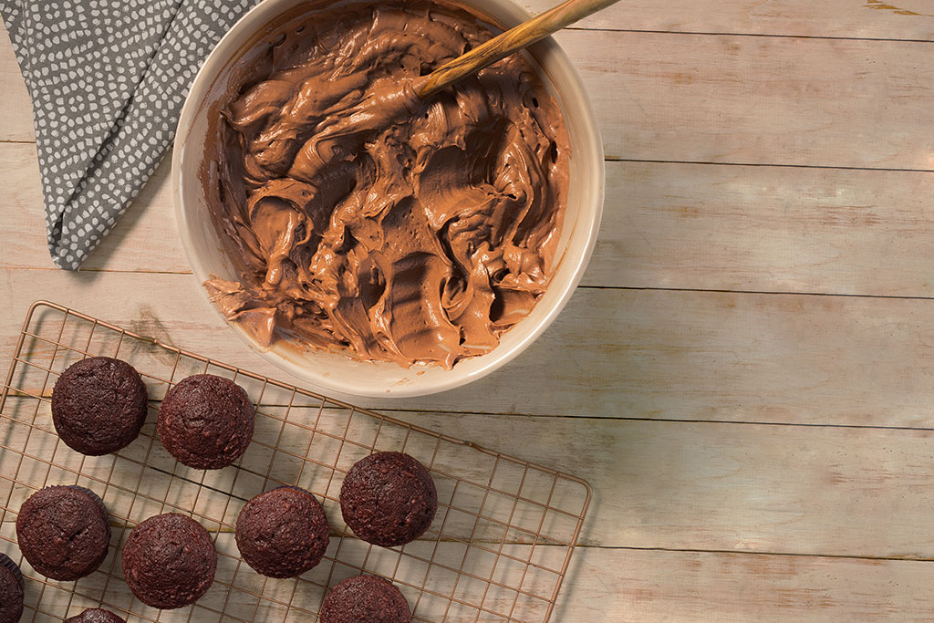 chocolate frosting in a bowl and chocolate brownies on a cooling rack on a wooden table. 