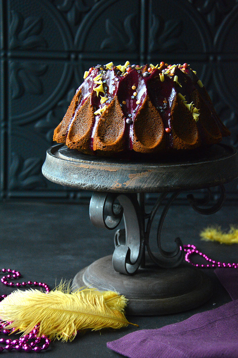 An intricate chocolate bundt cake with purple glaze and light yellow decor sits on an iron cake stand against a dark backdrop. Yellow feathers and purple beads lay underneath the cake. 